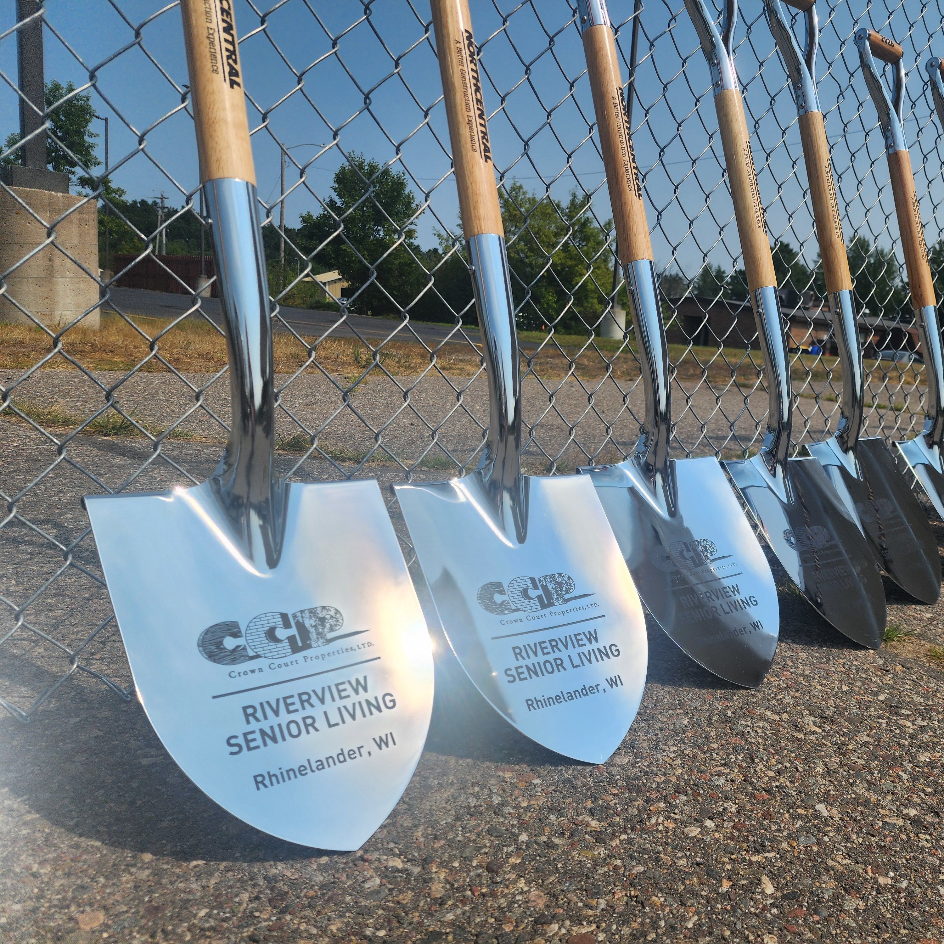 Close-up of ceremonial shovels with engraved blades lined up in front of a fence at an outdoor groundbreaking in Oneida County, Wisconsin