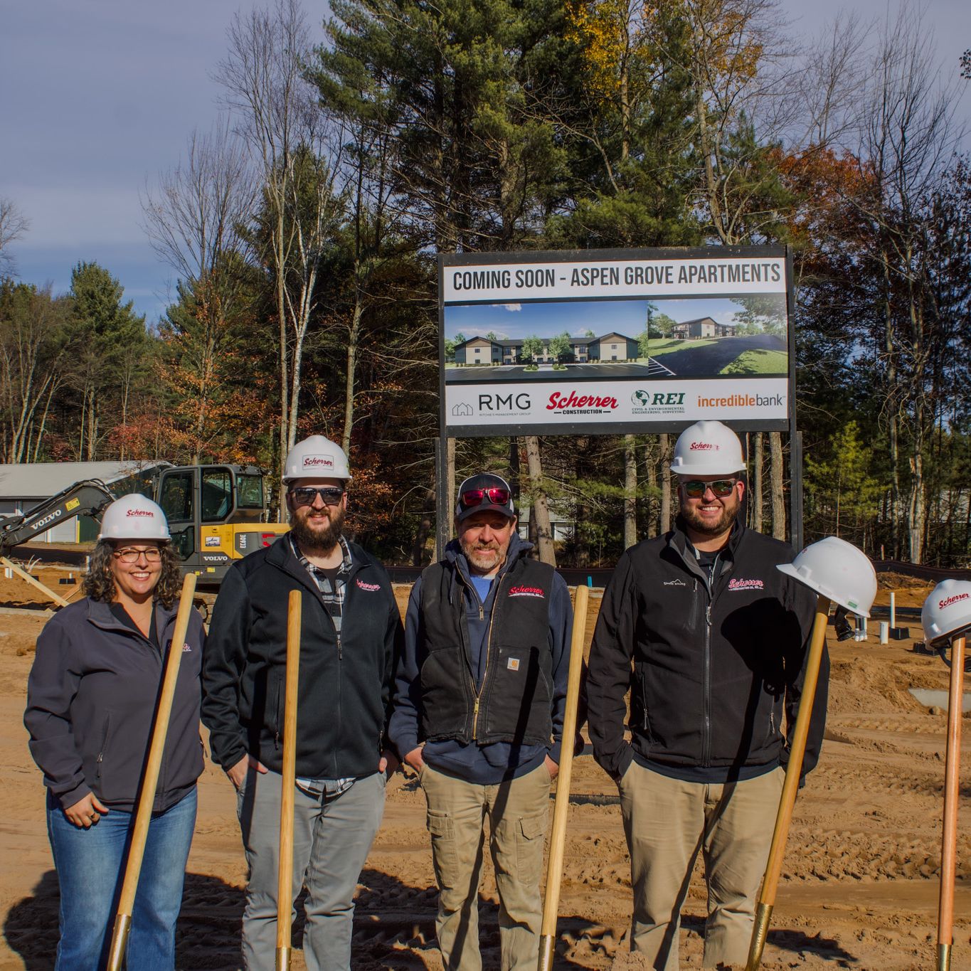 Four people wearing hard hats and holding shovels pose at a construction site in front of a large project sign and surrounded by trees