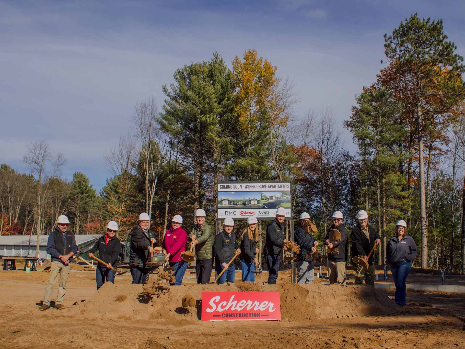 A group of people wearing hard hats participate in a groundbreaking ceremony on a construction site surrounded by fall trees in Oneida County, Wisconsin