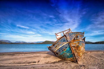 Aged and weathered boat resting on a sandy beach with bright blue skies and distant hills in the background