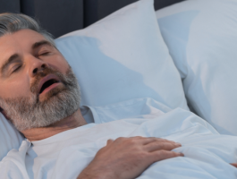 Mittelalter Mann mit grauem Bart schläft in Rückenlage im Bett mit geöffnetem Mund, Hinweise auf mögliche Schlafapnoe oder Schnarchen. Schlafzimmerumgebung mit weißen Kissen und Bettdecke, Fokus auf Atemprobleme im Schlaf.