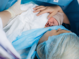 Frau liegt während eines Kaiserschnitts im Operationssaal, hält ihr Neugeborenes zum ersten Hautkontakt im Arm. Das Baby wird von medizinischem Personal unterstützt und liegt geschützt in Tüchern.