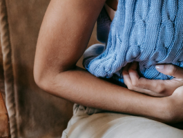 Person hält sich mit einem blauen Wärmekissen den Bauch, sitzt leicht nach vorne gebeugt und zeigt sichtbare Bauchschmerzen – symbolische Darstellung von krampfartigen Beschwerden wie Koliken oder starken Unterbauchschmerzen.
