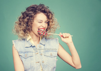 Eine Frau mit lockigem Haar in einer ärmellosen Jeansjacke hält einen großen, bunten Lollipop und beißt hinein, während sie ein Auge zwinkert. Der Hintergrund ist grün.