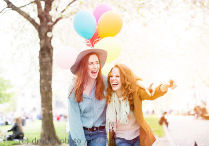Zwei lachende Frauen stehen im Freien unter einem blühenden Baum. Eine von ihnen trägt einen Hut und hält bunten Luftballons. Sie scheinen einen freudigen Moment zu teilen.