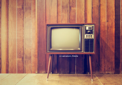 Ein retro Fernseher mit Holzgehäuse und vier Beinen steht vor einer Wand mit holzverkleideten Paneelen. Der Bildschirm des Fernsehers ist ausgeschaltet.