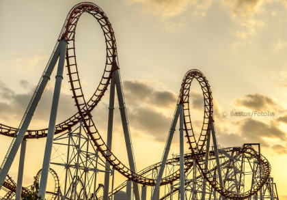 Das Bild zeigt eine Achterbahn mit mehreren Loopings und hohen Stützen vor einem Himmel bei Sonnenuntergang. Die Struktur der Achterbahn hebt sich deutlich gegen den orangefarbenen Himmel mit vereinzelten Wolken ab.