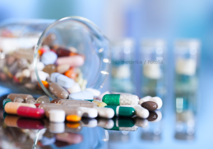 Ein umgestürztes Glasgefäß mit verschiedenen, bunten Medikamentenkapseln und -tabletten, die auf einer glänzenden Oberfläche verstreut sind. Im Hintergrund stehen unscharfe Glasfläschchen.