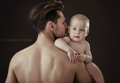 Ein Mann hält ein Baby auf dem Arm. Beide sind oberkörperfrei. Der Mann ist von hinten zu sehen, während das Baby nach vorne schaut. Der Hintergrund ist dunkel und schlicht.