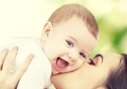 Eine Nahaufnahme einer Frau, die ein lächelndes Baby auf die Wange küsst. Das Baby hat helle Haut und blaue Augen und trägt ein weißes Outfit. Im Hintergrund sind unscharfe grüne & gelbe Farbtöne zu sehen, die ein Gefühl von Natur und Frische vermitteln.