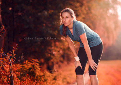 Eine Frau in Sportkleidung beugt sich nach dem Laufen nach vorne und legt ihre Hände auf die Knie. Sie sieht erschöpft aus und atmet schwer. Der Hintergrund zeigt eine herbstliche Waldlandschaft im warmen Licht der untergehenden Sonne.