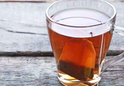 Eine Glas-Tasse mit heißem Tee und einem Teebeutel darin steht auf einer rustikalen, grauen Holzoberfläche.