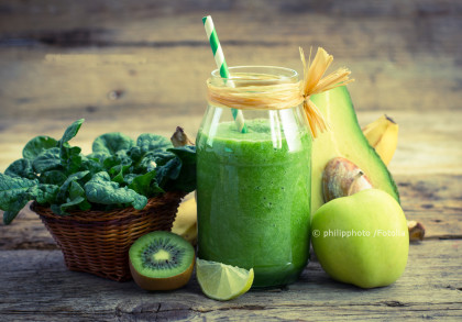 Grüner Smoothie in einem Glas mit Strohhalm, umgeben von frischem Spinat in einem Korb, einer halben Kiwi, einer Zitronenscheibe, einem Apfel und einer Avocado-Hälfte, die auf einem rustikalen Holzuntergrund arrangiert sind.