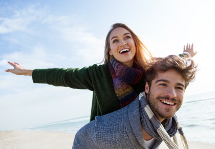 Ein lächelnder Mann trägt eine lächelnde Frau auf dem Rücken am Strand. Die Frau breitet ihre Arme aus, als ob sie fliegt. Beide tragen warme Kleidung und Schals. Der Himmel ist leicht bewölkt und das Meer ist im Hintergrund zu sehen.