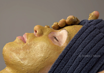 Eine Person liegt mit geschlossenen Augen, während eine goldfarbene Gesichtsmaske auf ihr Gesicht aufgetragen wird. Ihr Kopf ist mit einem dunkelblauen Handtuch umwickelt.