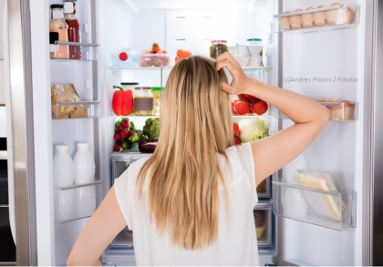 Eine Frau mit langen blonden Haaren steht vor einem offenen Kühlschrank, der mit verschiedenen Lebensmitteln gefüllt ist. Sie kratzt sich am Kopf und scheint darüber nachzudenken, was sie nehmen soll.