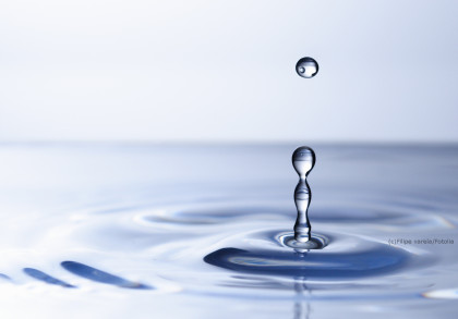 Tropfende Wassertropfen, die auf eine Wasseroberfläche fallen und konzentrische Kreise bilden. Die Oberfläche ist glatt und reflektierend, und der Hintergrund ist in weichen Blau- und Grautönen gehalten.