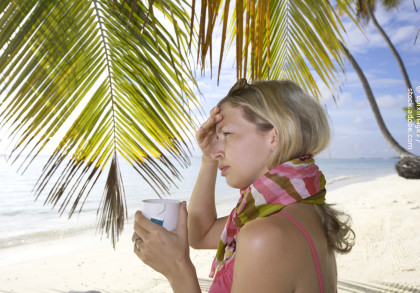 Eine Frau steht am Strand unter Palmen. Sie trägt ein ärmelloses Oberteil und einen bunten Schal. In ihrer rechten Hand hält sie eine Tasse und mit der linken Hand stützt sie ihre Stirn. Im Hintergrund ist der Ozean und ein klarer Himmel zu sehen.