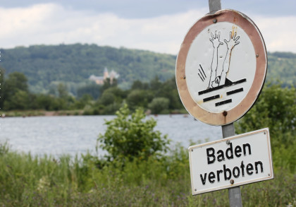 Ein Schild mit der Aufschrift "Baden verboten" und einem Symbol, das eine Person zeigt, die ertrinkt. Im Hintergrund sieht man einen See, umgeben von Bäumen und Hügeln.