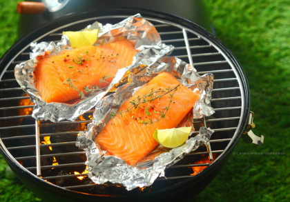 Zwei Lachsfilets liegen in Aluminiumfolie auf einem Grillrost. Sie sind mit frischen Kräutern und Gewürzen garniert. Eine Zitronenscheibe befindet sich auf jedem Lachsfilet. Der Grill steht draußen auf grünem Gras.