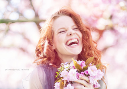 Eine Frau mit langen, lockigen, roten Haaren lacht herzlich und hält einen Strauß pinker Blumen. Der Hintergrund ist mit verschwommenen Pastellfarben gesättigt und deutet auf einen blühenden Garten oder Park hin.