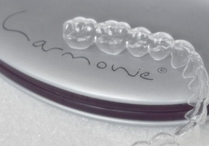 Durchsichtige Zahnspange auf einem silbernen Etui mit der Aufschrift "Harmonie". Das Etui liegt auf einer weißen Oberfläche.