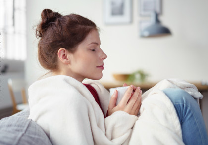 Eine Frau mit braunen Haaren sitzt gemütlich auf einem Sofa, in eine weiße Decke gehüllt, und hält eine Tasse in beiden Händen. Sie hat die Augen geschlossen und wirkt entspannt. Im Hintergrund ist unscharf eine Zimmerdekoration zu erkennen.