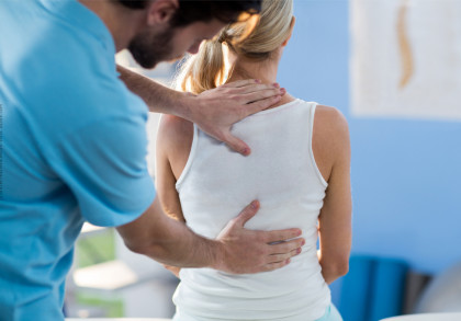 Ein Physiotherapeut behandelt eine Frau, indem er ihr mit seinen Händen den oberen und unteren Rücken massiert. Die Frau sitzt mit dem Rücken zur Kamera und trägt ein weißes Oberteil. Der Therapeut trägt ein hellblaues Hemd.