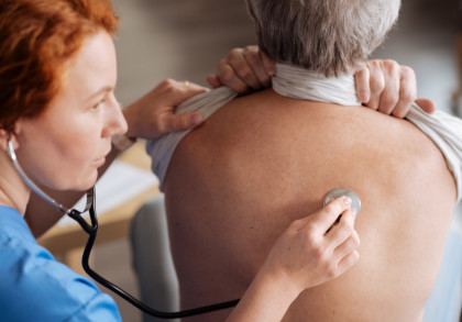 Eine Ärztin hört mit einem Stethoskop den Rücken eines älteren Patienten ab. Der Patient sitzt mit freiem Oberkörper und die Ärztin ist konzentriert bei der Arbeit.