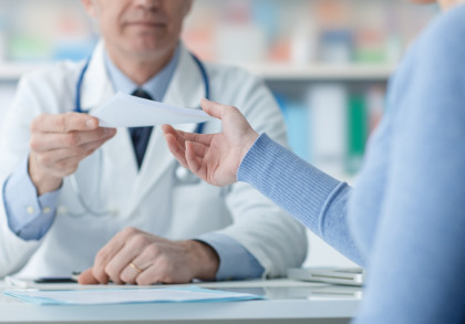 Ein Arzt in einem weißen Kittel überreicht einem Patienten ein Rezept. Der Patient trägt einen blauen Pullover. Beide sitzen an einem Schreibtisch in einem gut beleuchteten Büro, das mit Ordnern und medizinischen Geräten im Hintergrund ausgestattet ist.