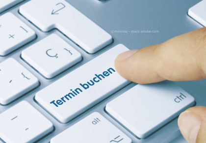 Eine Nahaufnahme einer Computer-Tastatur mit einer Taste beschriftet als "Termin buchen". Ein Finger drückt diese Taste.