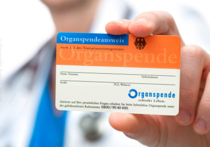 Ein Hand hält einen deutschen Organspendeausweis. Der Ausweis ist orange und blau und hat Felder für Name, Vorname, Geburtsdatum, Straße, Postleitzahl und Wohnort. Oben steht "Organspendeausweis nach § 2 des Transplantationsgesetzes".