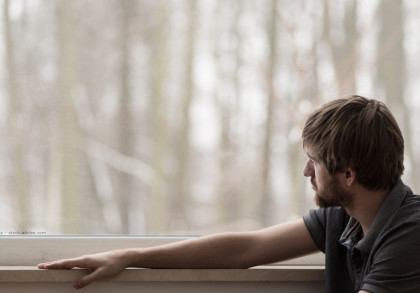 Ein Mann mit kurzen braunen Haaren und Bart sitzt am Fenster und schaut nach draußen. Seine linke Hand ruht auf der Fensterbank. Draußen sind Bäume und ein unscharfer Hintergrund zu sehen. Die Stimmungist  kalt, möglicherweise winterliche Stimmung.