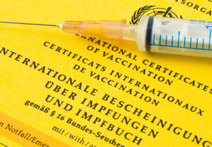 Ein gelbes internationales Zertifikat über Impfungen und Impfbuch liegt auf einer Oberfläche. Darauf liegt eine Einwegspritze. Der Titel des Dokuments ist in mehreren Sprachen, darunter Deutsch, Englisch und Französisch, geschrieben.