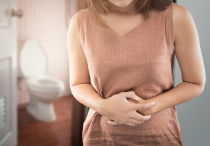Eine Frau steht vor einem Badezimmer mit einem leicht geöffneten Tür. Im Hintergrund ist eine Toilette sichtbar. Die Frau trägt ein ärmelloses, braunes Kleid und hält sich den Bauch, was auf Bauchschmerzen hinweisen könnte.