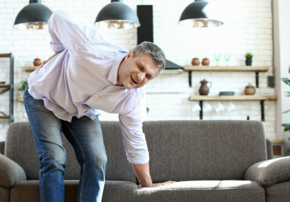 Ein Mann mittleren Alters mit grauen Haaren und einem hellen Hemd steht vor einem Sofa in einem modernen Wohnzimmer. Er hält sich mit schmerzerfülltem Gesichtsausdruck den unteren Rücken und stützt sich mit einer Hand auf das Sofa.