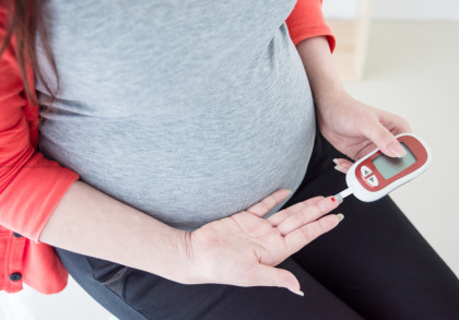 Eine schwangere Frau in grauem T-Shirt und rotem Pullover misst ihren Blutzucker mit einem Glukometer, indem sie einen Bluttropfen von ihrem Finger nimmt.