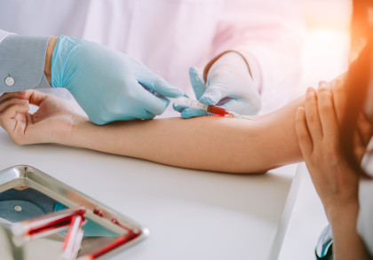 Eine Person erhält eine Blutabnahme in einem medizinischen Umfeld. Eine zweite Person mit Einweghandschuhen zieht Blut mit einer Spritze aus der Vene im Arm der ersten Person. Im Vordergrund steht ein Metallschale mit Blutprobenröhrchen.