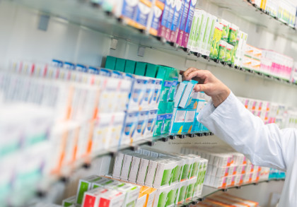 Ein Apotheker oder eine Apothekerin in einem weißen Kittel greift nach einer Medikamentenverpackung aus einem Regal in einer Apotheke. Das Regal ist gefüllt mit verschiedenen bunten Medikamentenschachteln in unterschiedlichen Größen und Farben.