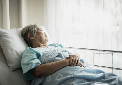 Ältere Frau liegt in einem Krankenhausbett und blickt nachdenklich aus dem Fenster. Sie trägt ein blaues Krankenhaushemd und ist mit einer blauen Decke zugedeckt. Licht strömt durch die weißen Vorhänge.