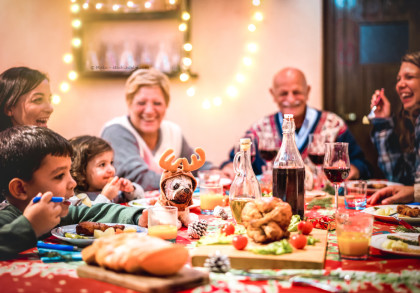 Eine Gruppe von Menschen sitzt an einem festlich gedeckten Tisch und lächelt, während sie eine Mahlzeit genießen. Der Tisch ist mit verschiedenen Speisen, Getränken und Weihnachtsdekorationen geschmückt.