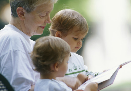 Eine ältere Frau sitzt draußen auf einer Bank und liest zwei kleinen Kindern ein Buch vor. Alle drei tragen weiße T-Shirts und schauen konzentriert auf das Buch. Die Umgebung ist leicht verschwommen, was auf einen Garten oder Park schließen lässt.