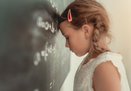 Ein kleines Mädchen mit blonden Zöpfen in einem weißen Spitzenkleid lehnt ihren Kopf gegen eine Tafel, auf der mathematische Formeln stehen. Sie wirkt entmutigt oder frustriert.
