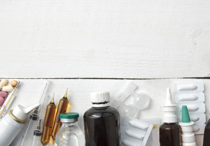 Medikamente und medizinische Geräte auf einem weißen Holzuntergrund. Es gibt verschiedene Tabletten, Sirupflaschen, Nasensprays, Ampullen, eine Spritze, ein Fieberthermometer, ein digitales Blutdruckmessgerät und eine Gesichtsmaske.