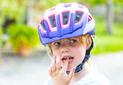 Ein kleines Kind trägt einen lila-rosa Fahrradhelm und steckt sich einen Finger in den Mund. Der Hintergrund ist verschwommen und zeigt grüne Pflanzen.