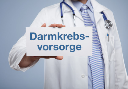 Ein Arzt in einem weißen Kittel und einer gestreiften Krawatte hält ein Schild mit der Aufschrift "Darmkrebsvorsorge". Ein Stethoskop hängt um seinen Hals.