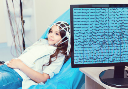Ein Kind liegt auf einer Liege und trägt eine Mütze mit Elektroden, die an mehrere Kabel angeschlossen sind. Auf einem Bildschirm neben dem Kind ist eine EEG-Aufzeichnung der Gehirnaktivität zu sehen.