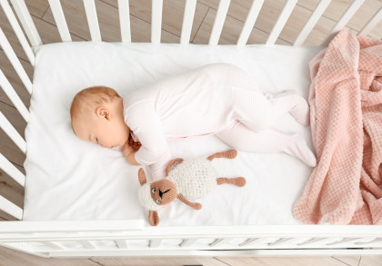 Ein Baby in weißem Strampler schläft auf der Seite in einem weißen Kinderbett. Neben dem Baby liegt ein gestricktes Stofftier in Form eines Schafes. Eine rosa Decke befindet sich am Rand des Kinderbettes.