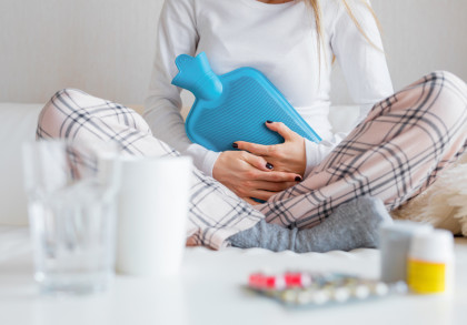 Eine Person sitzt auf einem Sofa und hält eine blaue Wärmflasche auf ihrem Bauch. Sie trägt ein weißes Langarmshirt und karierte Pyjamahosen. Im Vordergrund sind ein Glas Wasser und einige Medikamente zu sehen.