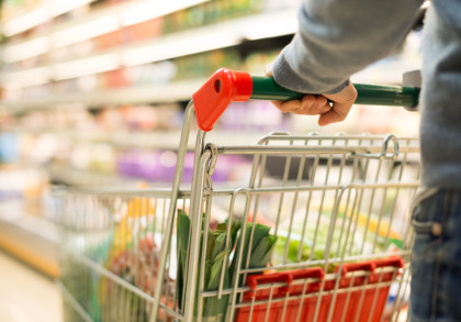 Eine Person hält einen Einkaufswagen im Supermarkt. Der Wagen ist teilweise gefüllt mit verschiedenen Lebensmitteln. Im Hintergrund sind verschwommene Regale mit weiteren Produkten zu sehen.